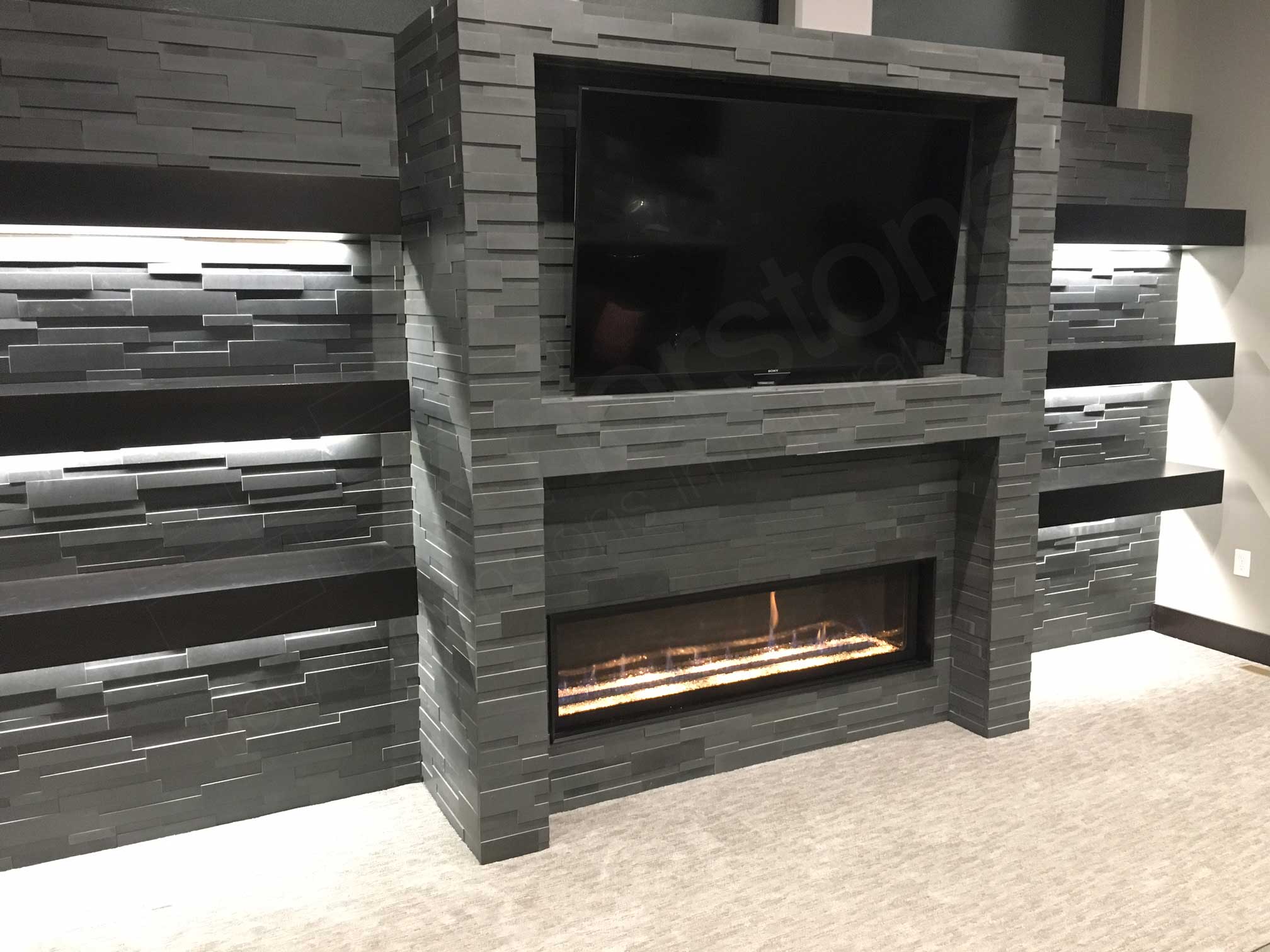 Gas fireplace and big screen tv recessed into a stone wall clad in grey basalt 3D ledge stone flanked on either side by floating shelves